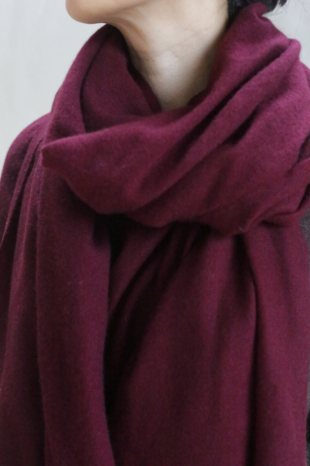 A person with short dark hair wears the Tissue Weight Cashmere Scarf in Garnet, wrapped snugly around their neck and shoulders. Only the lower half of their face and part of their upper torso are visible.