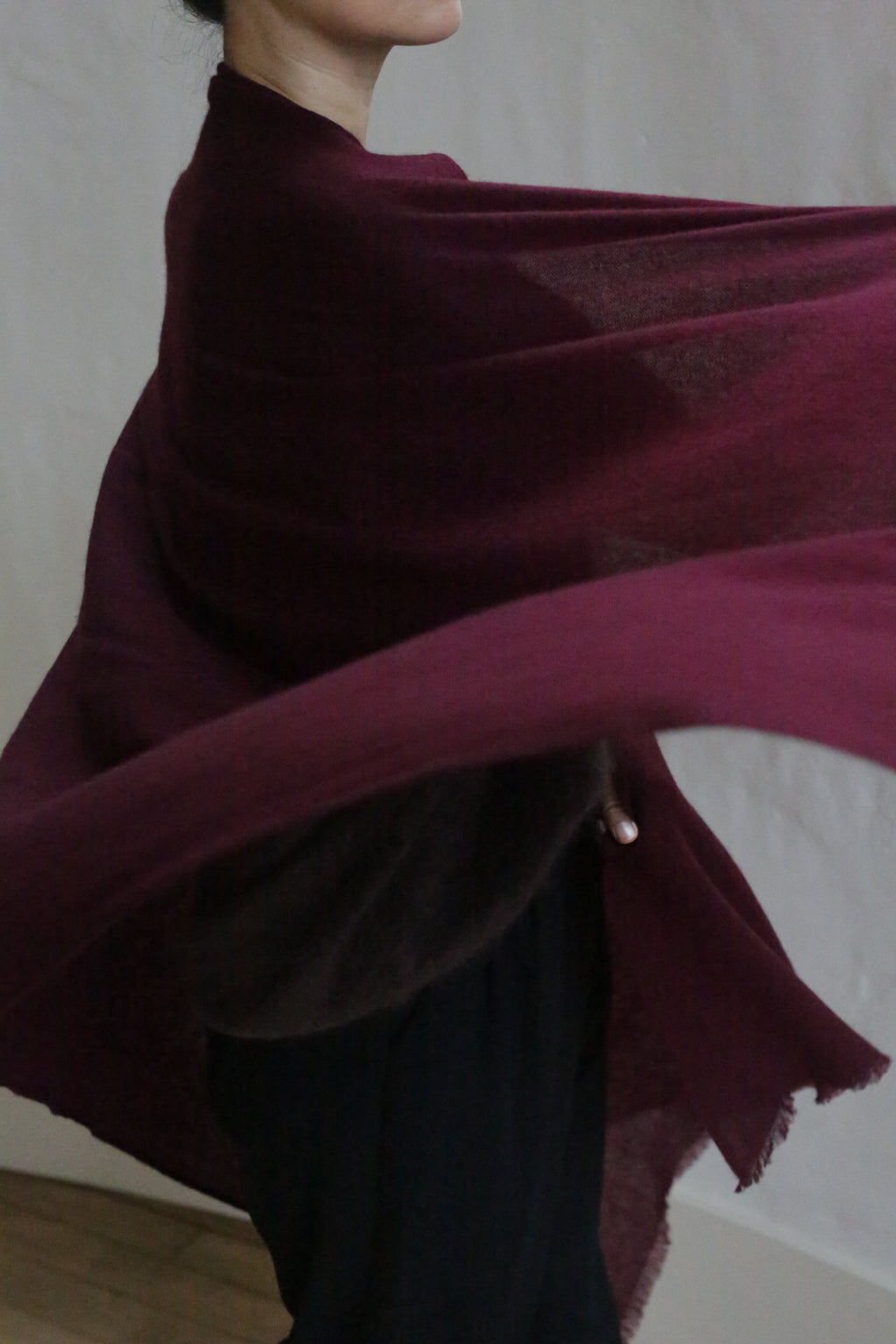 A person with short dark hair drapes the Garnet Tissue Weight Cashmere Scarf around their shoulders, its 100% cashmere fabric spilling outward against a plain light background, with their face partially out of frame.