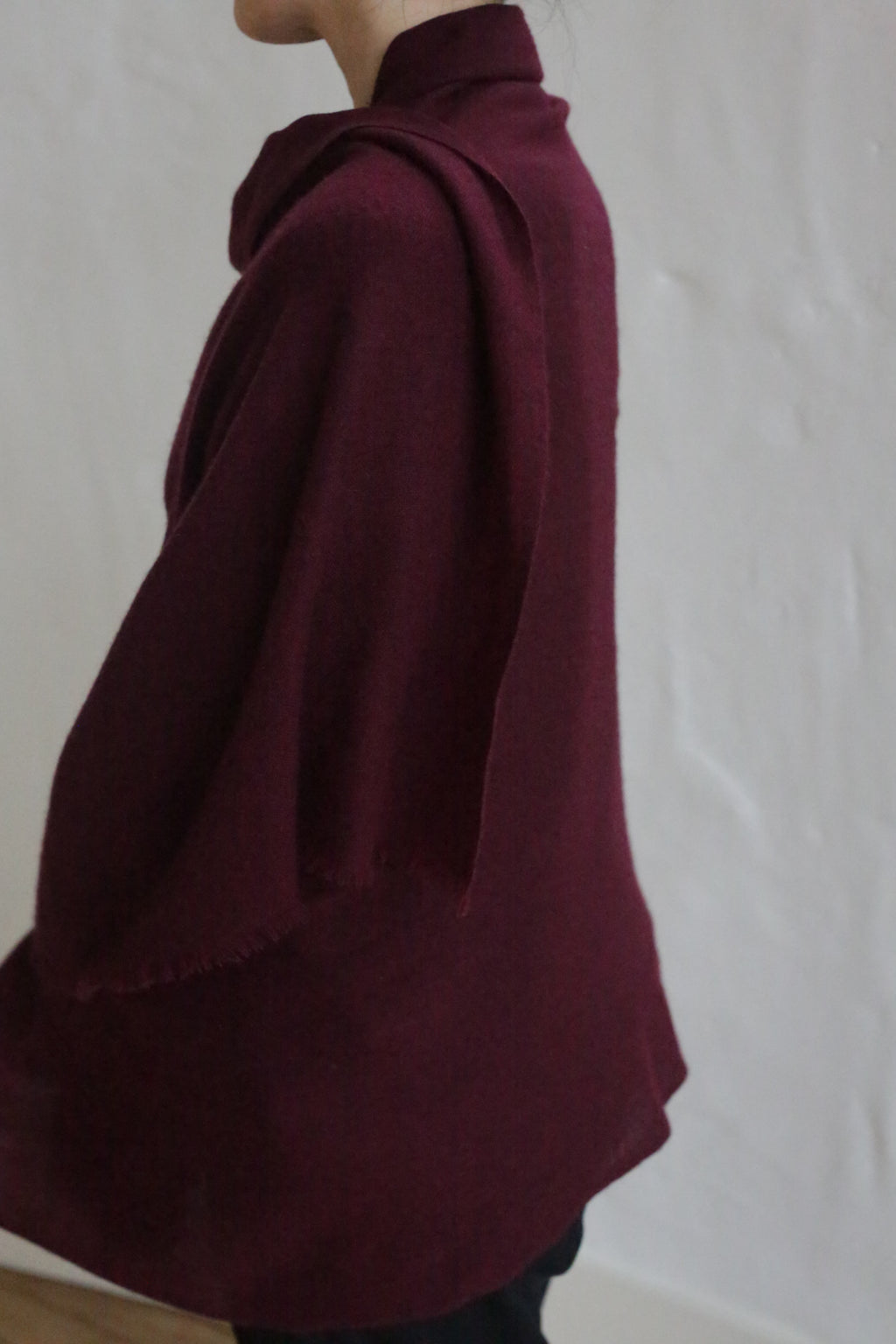 A person stands sideways against a white textured wall, wearing the Tissue Weight Cashmere Scarf in Garnet draped elegantly over their shoulders and arms. Only their upper body is visible, showcasing the luxurious cashmere.