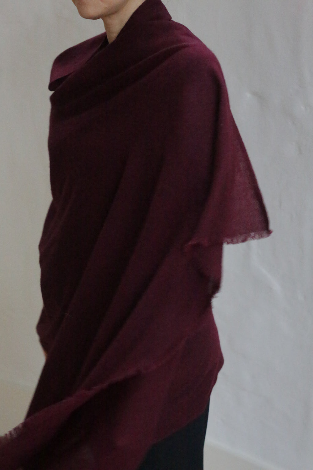 A person drapes the Tissue Weight Cashmere Scarf in Garnet over their shoulders, standing against a plain light background with their face partially out of view.