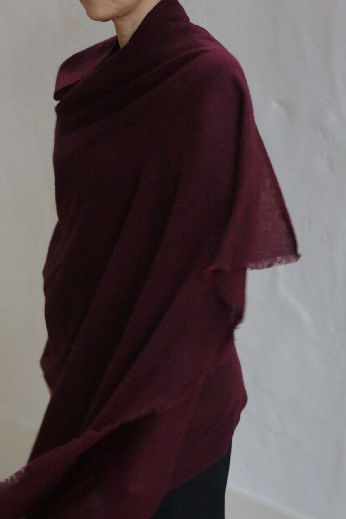 A person drapes the Tissue Weight Cashmere Scarf in Garnet over their shoulders, standing against a plain light background with their face partially out of view.