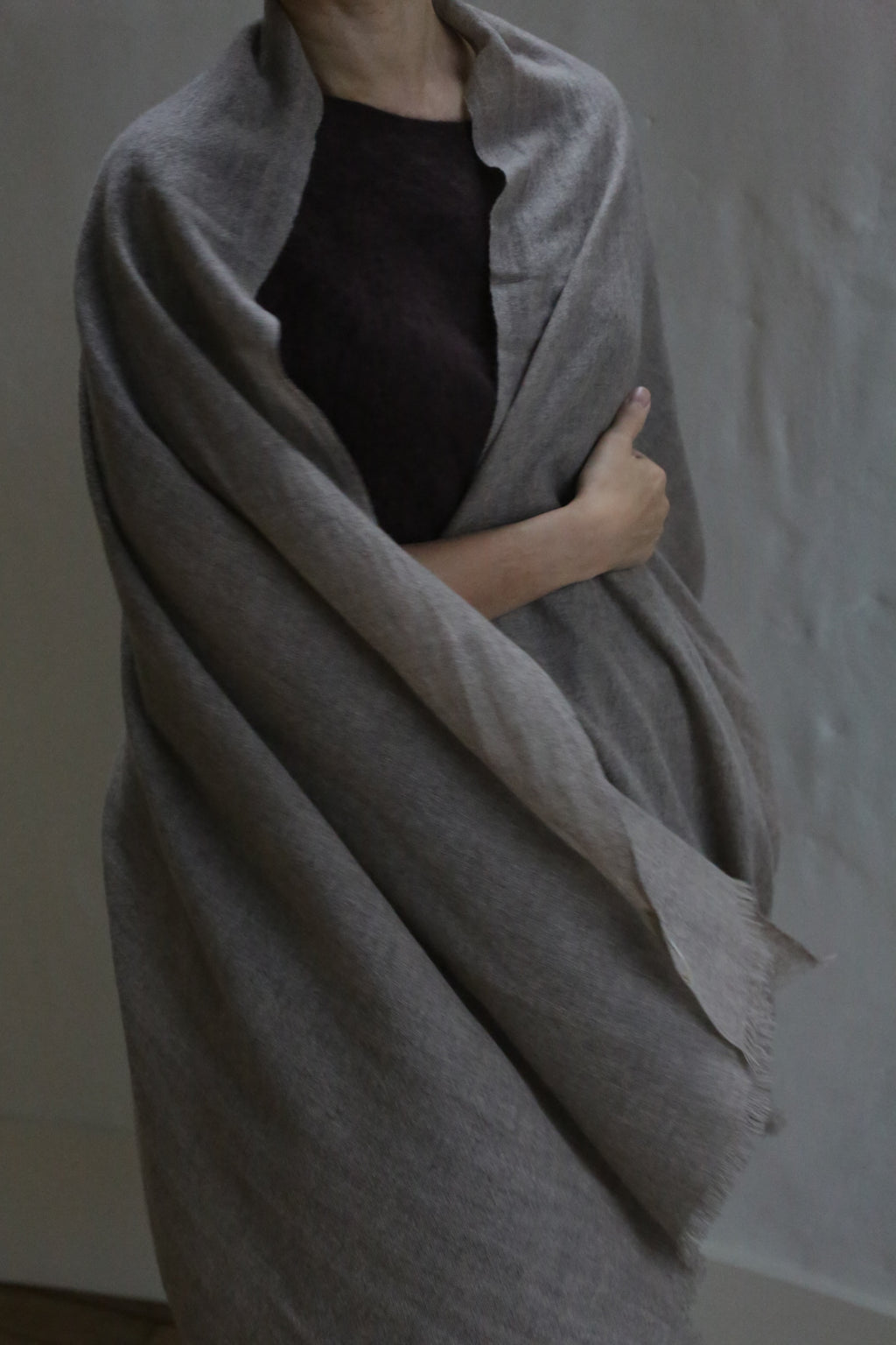 A person with light skin wears the Tissue Weight Cashmere Scarf in Natural, a large, light gray woven accessory. They hold it closed with one arm over a dark top. Their face is partly hidden against a plain, softly lit background.