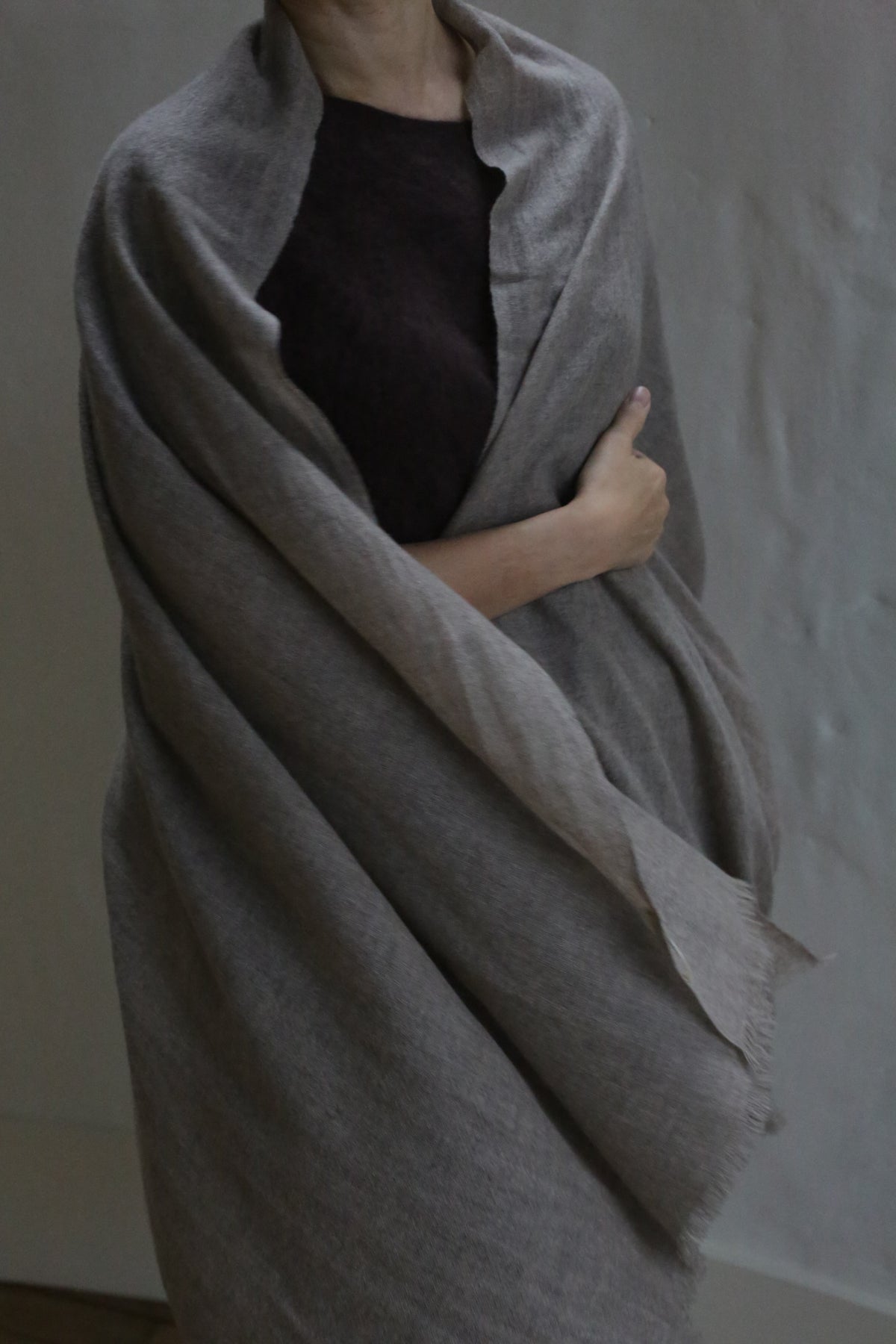 A person with light skin wears the Tissue Weight Cashmere Scarf in Natural, a large, light gray woven accessory. They hold it closed with one arm over a dark top. Their face is partly hidden against a plain, softly lit background.