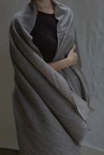 Load image into Gallery viewer, A person with light skin wears the Tissue Weight Cashmere Scarf in Natural, a large, light gray woven accessory. They hold it closed with one arm over a dark top. Their face is partly hidden against a plain, softly lit background.