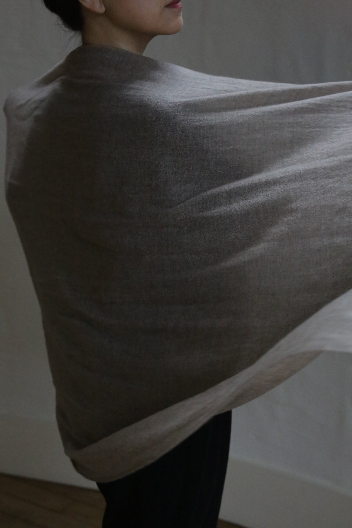 A person drapes the Tissue Weight Cashmere Scarf in Natural—a large, soft, grayish-brown 100% cashmere wrap—over their shoulders. Soft light highlights its luxurious texture and their side profile against a neutral background.