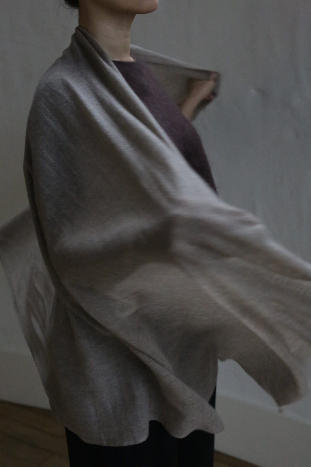 A person with dark hair, partially visible, is draped in the Tissue Weight Cashmere Scarf | Natural over a dark outfit. The lightweight 100% cashmere scarf flows gracefully, adding gentle movement against a softly lit, plain background.