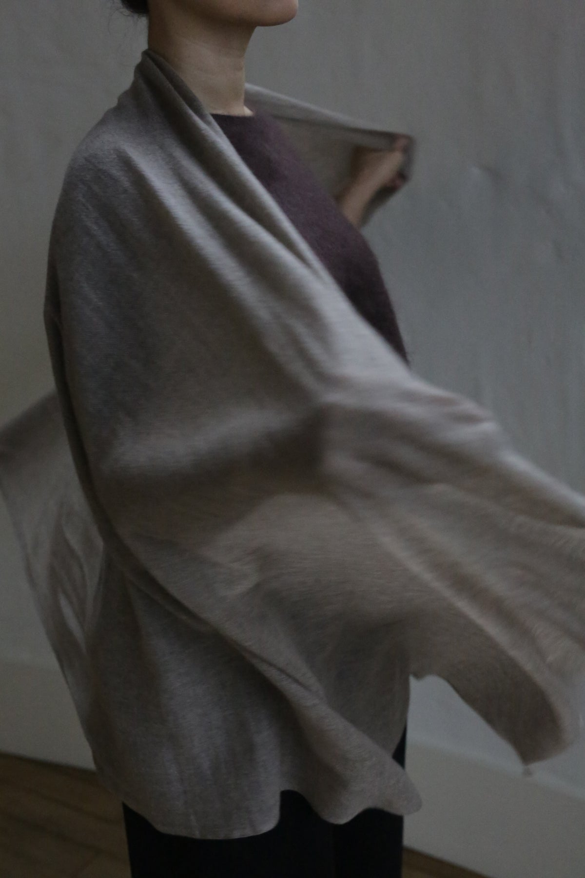 A person with dark hair, partially visible, is draped in the Tissue Weight Cashmere Scarf | Natural over a dark outfit. The lightweight 100% cashmere scarf flows gracefully, adding gentle movement against a softly lit, plain background.