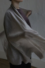 Load image into Gallery viewer, A person with dark hair, partially visible, is draped in the Tissue Weight Cashmere Scarf | Natural over a dark outfit. The lightweight 100% cashmere scarf flows gracefully, adding gentle movement against a softly lit, plain background.