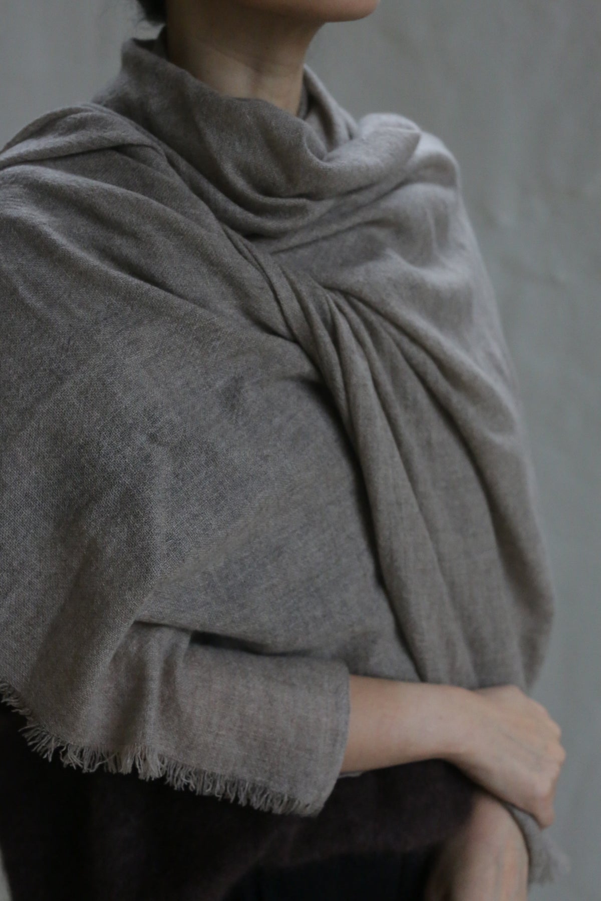 A person with light skin is shown from the shoulders down, wrapped in the Tissue Weight Cashmere Scarf in Natural—a soft, light brown woven accessory—across their shoulders and torso. One arm rests gently across their body against a neutral background.