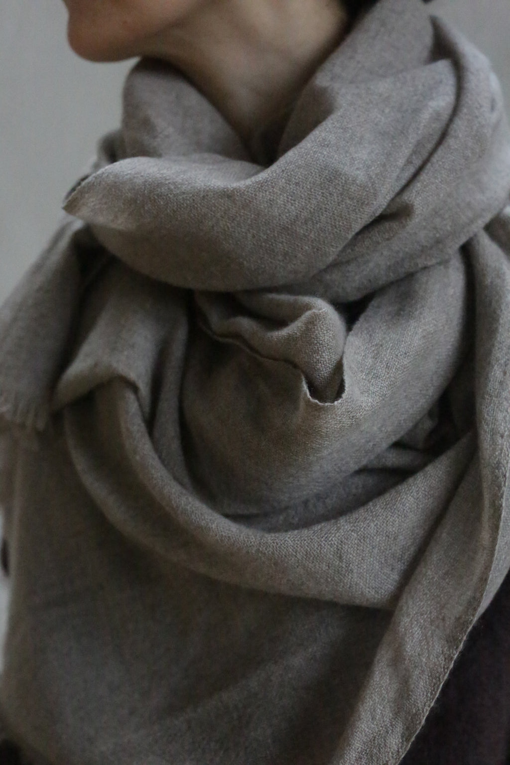 A close-up shows a person wearing the Tissue Weight Cashmere Scarf in Natural—a large, soft, grayish-brown 100% cashmere scarf—wrapped snugly around their neck and shoulders against a neutral background. The person's face is mostly out of frame.