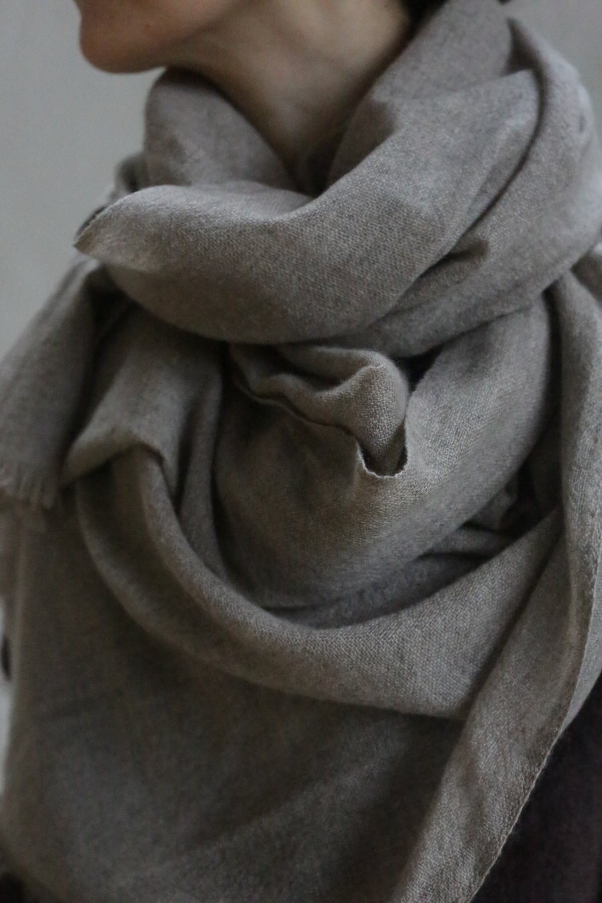 A close-up shows a person wearing the Tissue Weight Cashmere Scarf in Natural—a large, soft, grayish-brown 100% cashmere scarf—wrapped snugly around their neck and shoulders against a neutral background. The person's face is mostly out of frame.