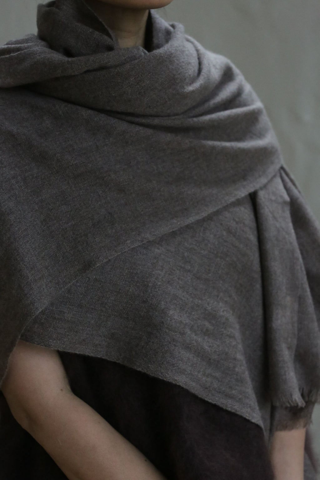 A person wears a Tissue Weight Cashmere Scarf in taupe, the soft woven fabric draped loosely around their shoulders. Their face is partially visible against a plain, neutral background.