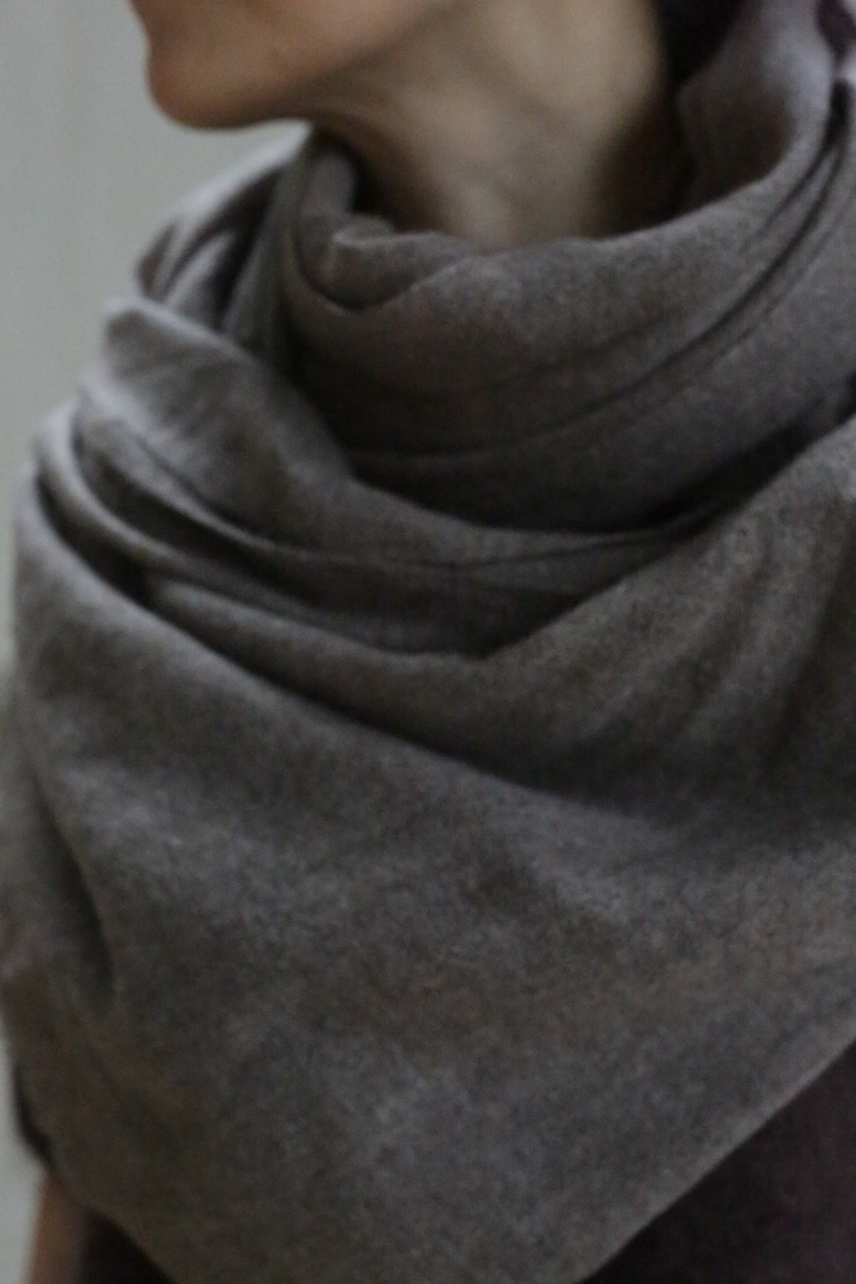 A close-up shows a person’s neck and shoulders wrapped in the Tissue Weight Cashmere Scarf in Taupe (36" x 82"). The face is partially out of frame, with a blurred, neutral background.