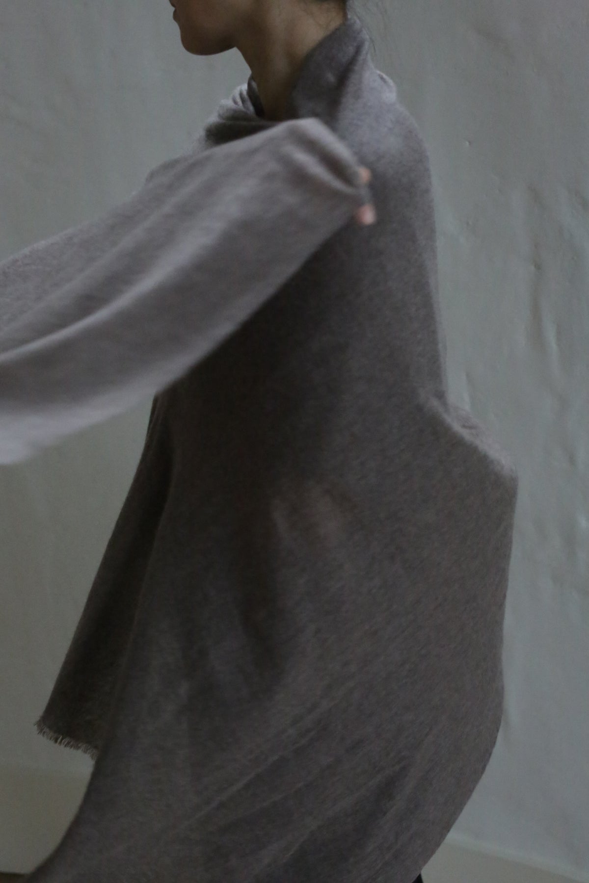 A person wears the Tissue Weight Cashmere Scarf in taupe, loosely draped with one arm extended, partially obscured against a plain light background. Their face is mostly out of frame.