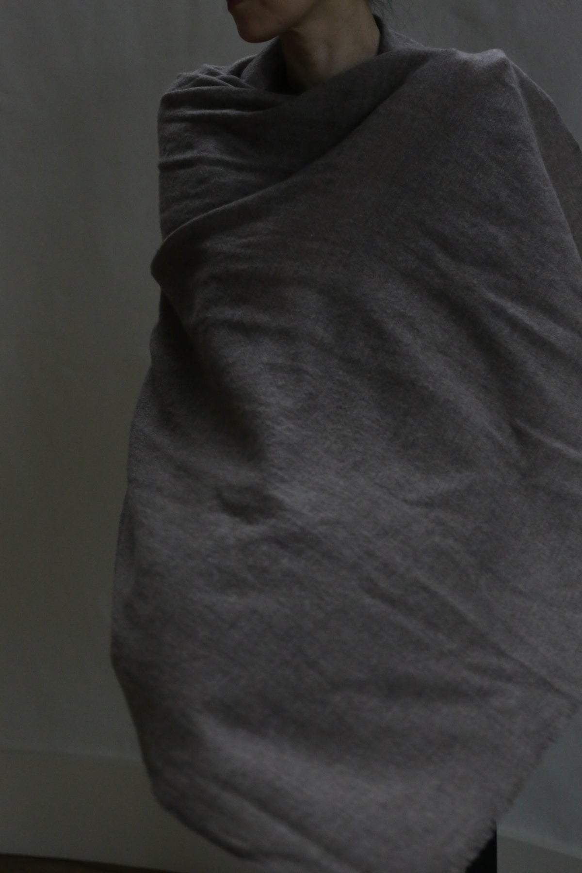 A person partially visible, wrapped in the Tissue Weight Cashmere Scarf in Taupe (36" x 82"), soft and greyish-brown, stands against a plain, dimly lit background with their face not fully shown.