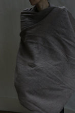 Load image into Gallery viewer, A person partially visible, wrapped in the Tissue Weight Cashmere Scarf in Taupe (36&quot; x 82&quot;), soft and greyish-brown, stands against a plain, dimly lit background with their face not fully shown.