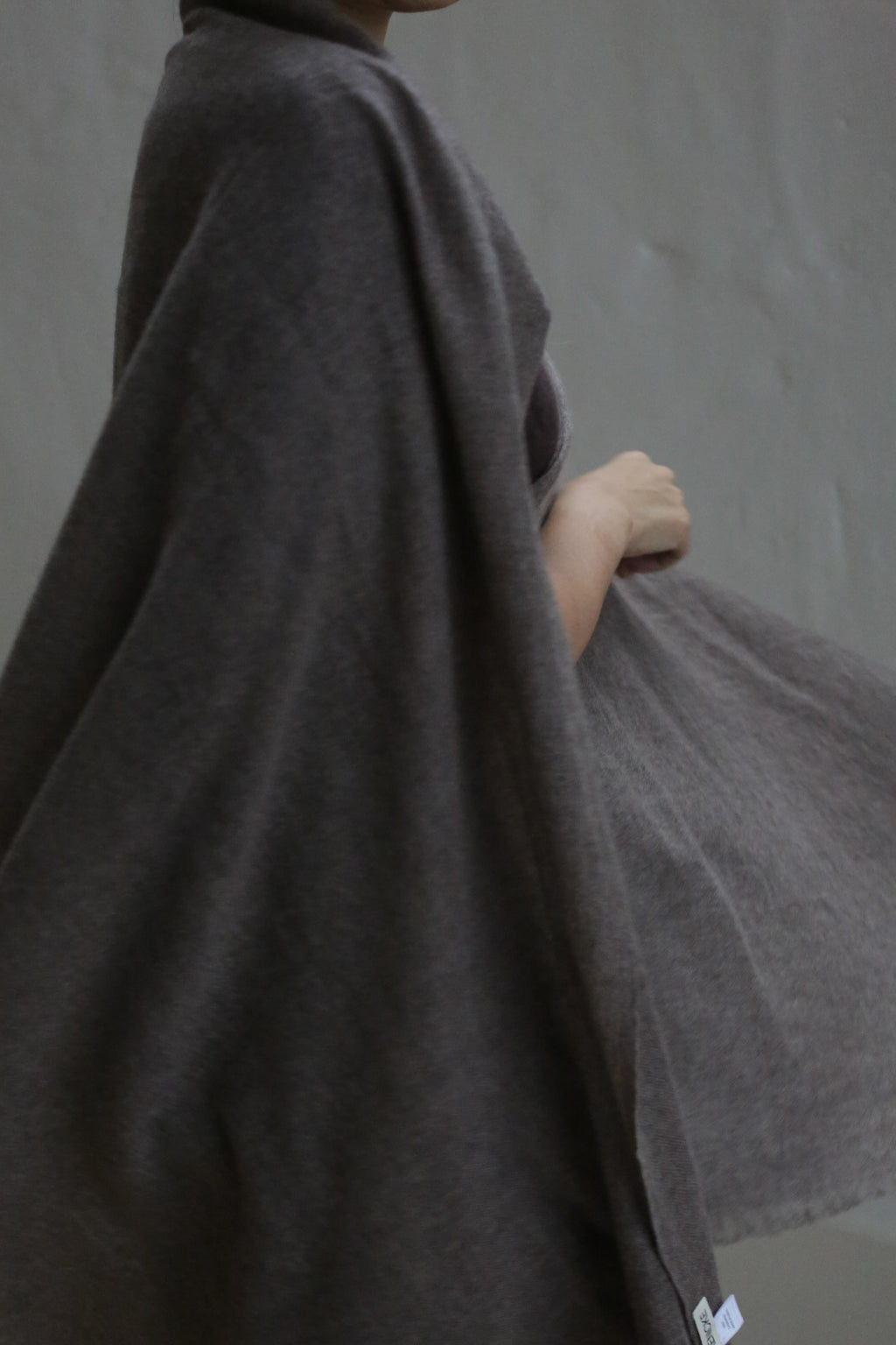 A person wears the Tissue Weight Cashmere Scarf in taupe, draped softly around their shoulders against a plain gray background, standing partially turned with one arm bent. Their face is mostly out of frame.