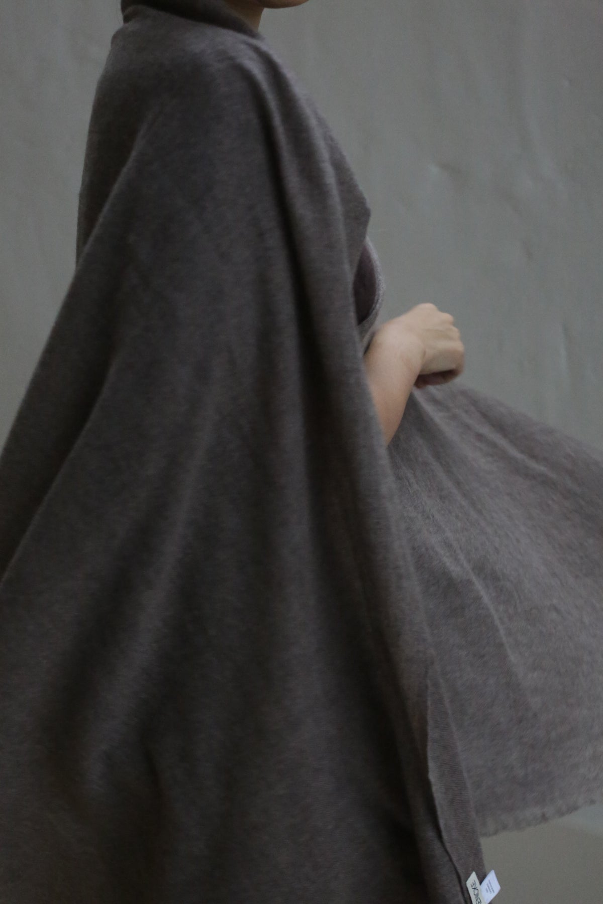 A person wears the Tissue Weight Cashmere Scarf in taupe, draped softly around their shoulders against a plain gray background, standing partially turned with one arm bent. Their face is mostly out of frame.