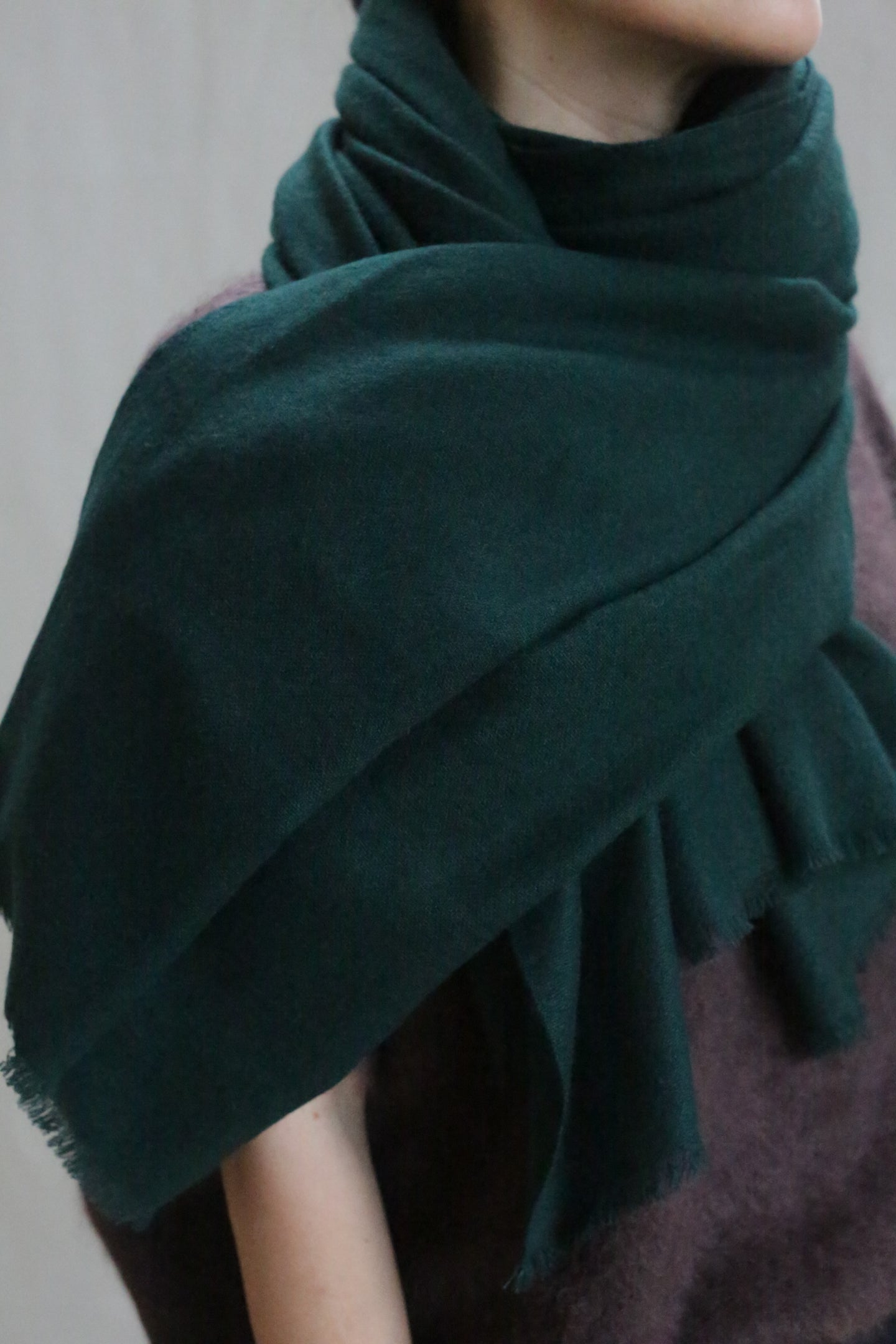 A person wears the Tissue Weight Cashmere Scarf in Forest (36 x 82), wrapped around their neck and shoulders over a purple sweater. The soft, woven cashmere looks warm, with the person's face partially out of frame.