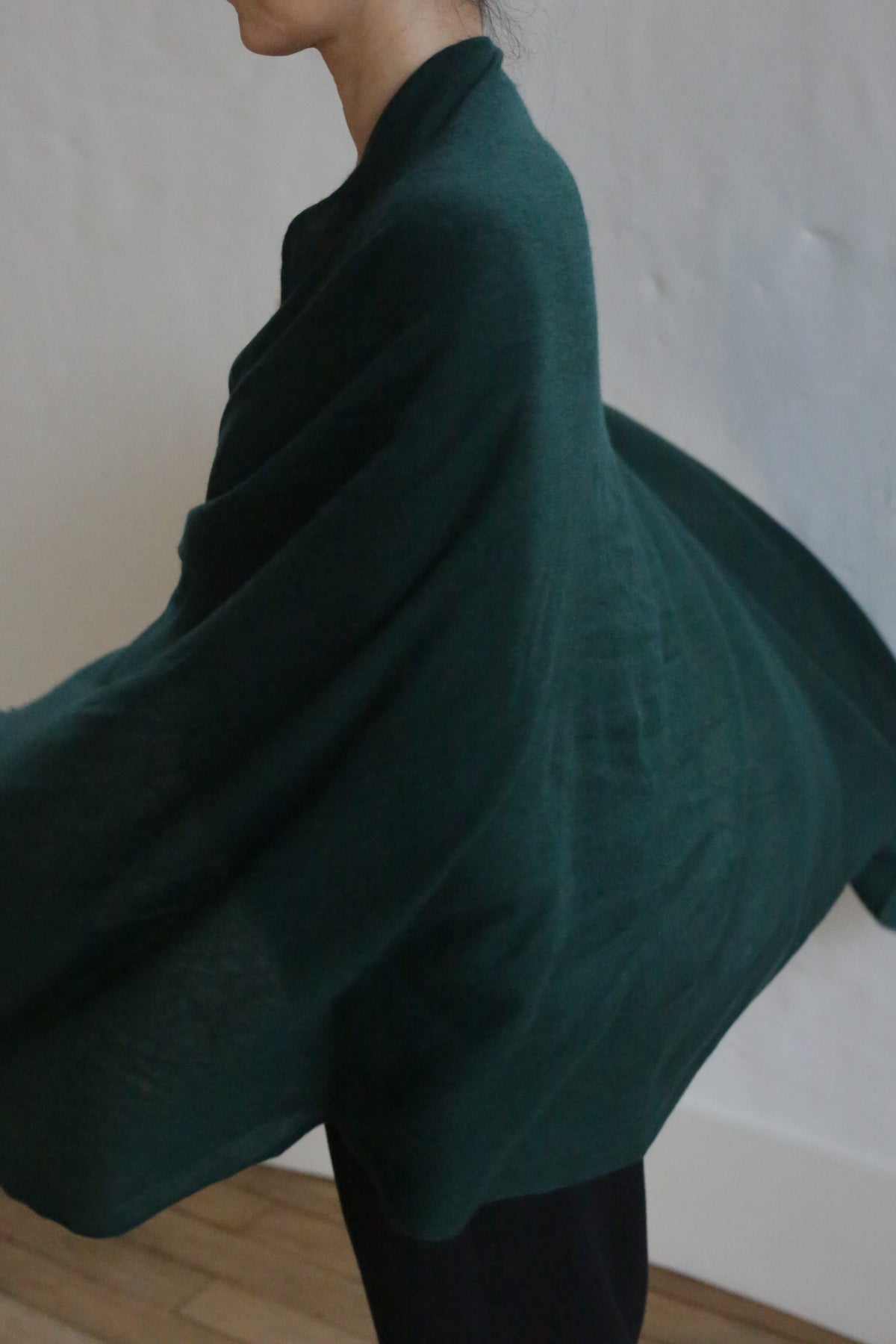 A person with short hair drapes the Tissue Weight Cashmere Scarf | Forest, measuring 36 x 82 inches, around their shoulders. The dark green scarf billows outward against a white wall and light wooden floor; their face is partially out of frame.