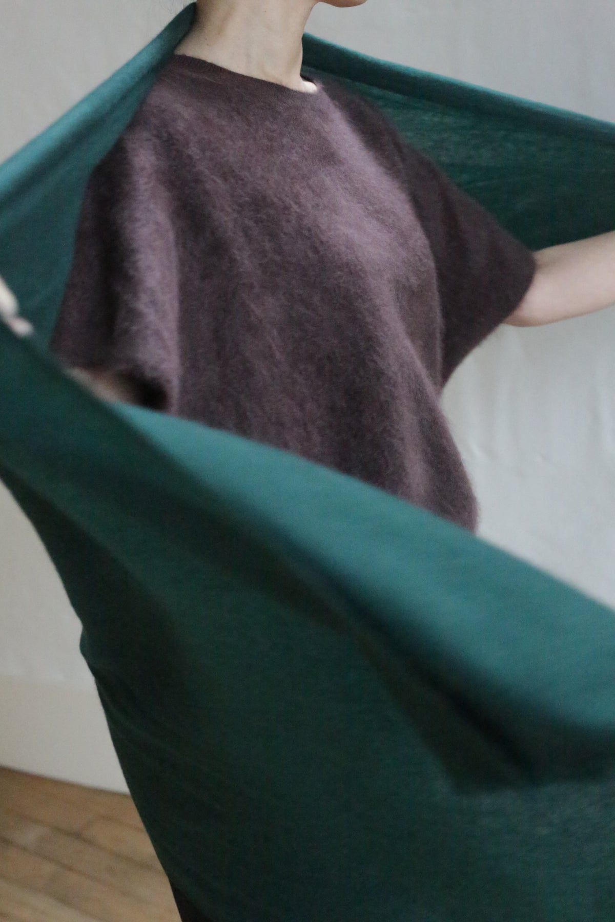 A person in a dark brown short-sleeve top stands partly hidden behind the Tissue Weight Cashmere Scarf in Forest, sized 36 x 82, which is draped diagonally. The softly lit background is plain.