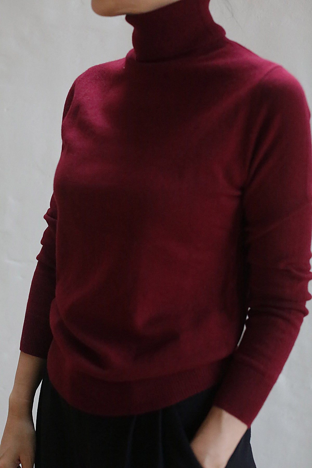 A person wears the Cashmere Turtleneck | Wine, its rich burgundy hue contrasting against a plain, light background. Their face is partially out of frame, with one hand resting loosely by their side.