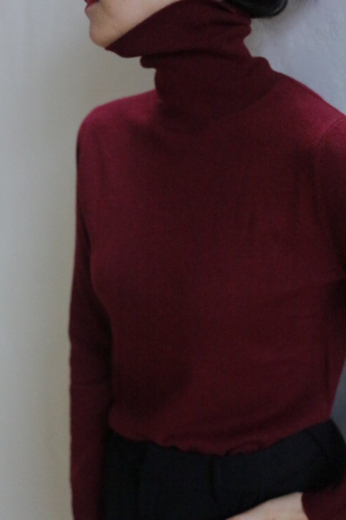 A person in the Cashmere Turtleneck | Wine, a fitted, long-sleeved dark red sweater, stands against a light wall with their head slightly turned and face partially out of frame.