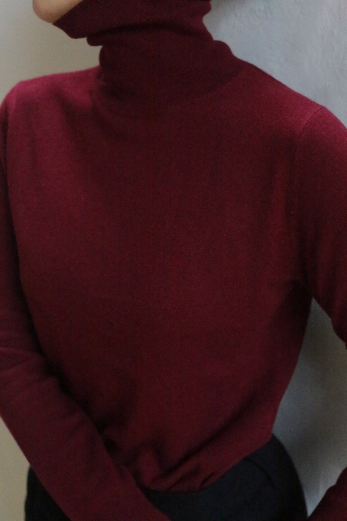 A person wearing the Cashmere Turtleneck | Wine, shown from shoulders to waist with black pants and their face partially out of frame. This 100% cashmere sweater is hand washable for easy care.