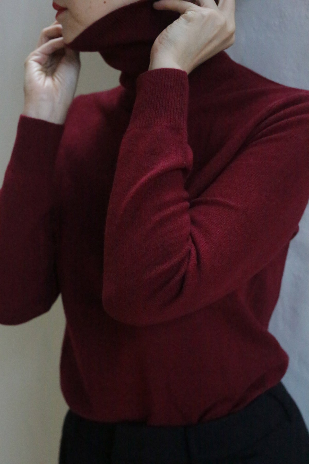 A person in the Cashmere Turtleneck | Wine is adjusting the collar with both hands. The cropped image displays only the upper body and part of the face.