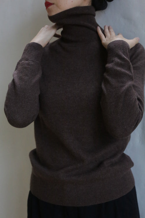 A person wearing the Cashmere Turtleneck | Coffee pulls the collar up to cover most of their face, with hands adjusting the neck. The background is plain and light-colored.