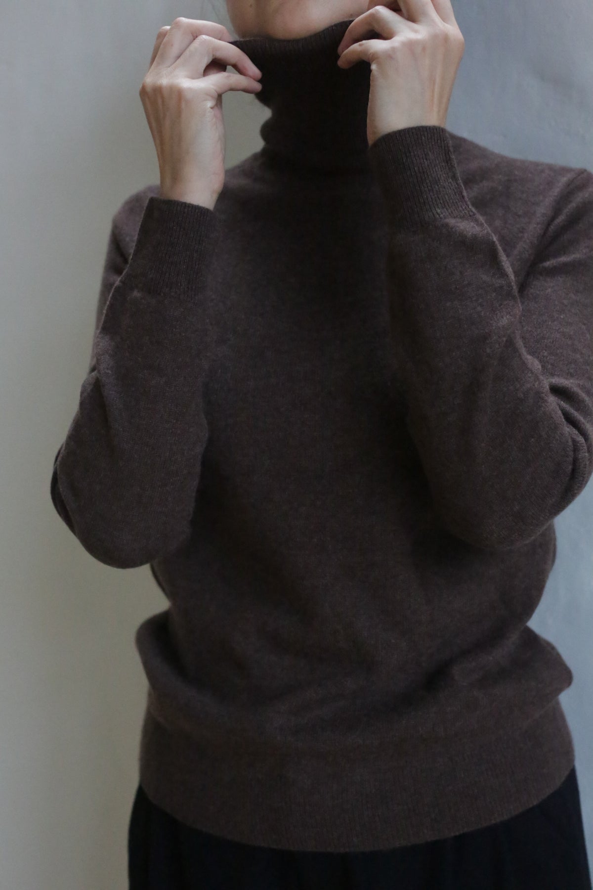 A person adjusts the collar of a hand washable Cashmere Turtleneck | Coffee, partially covering their face. The background is plain and light-colored, with most of their head out of frame.