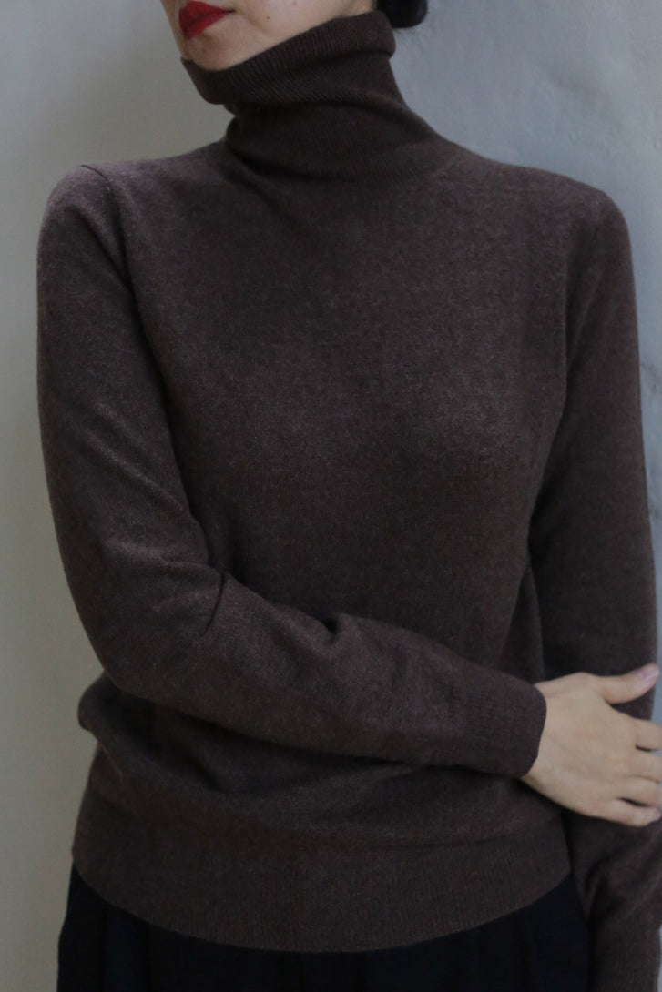 A person wears the Cashmere Turtleneck | Coffee, standing against a light-colored wall with one arm crossed and red lipstick partially visible.