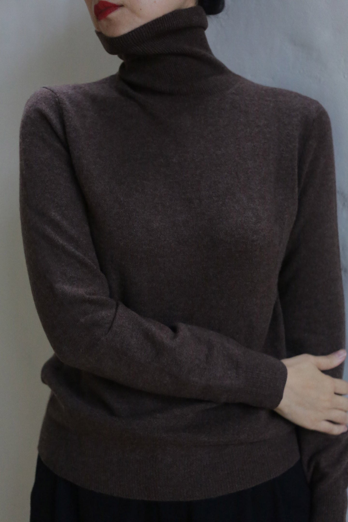 A person wears the Cashmere Turtleneck | Coffee, standing against a light-colored wall with one arm crossed and red lipstick partially visible.