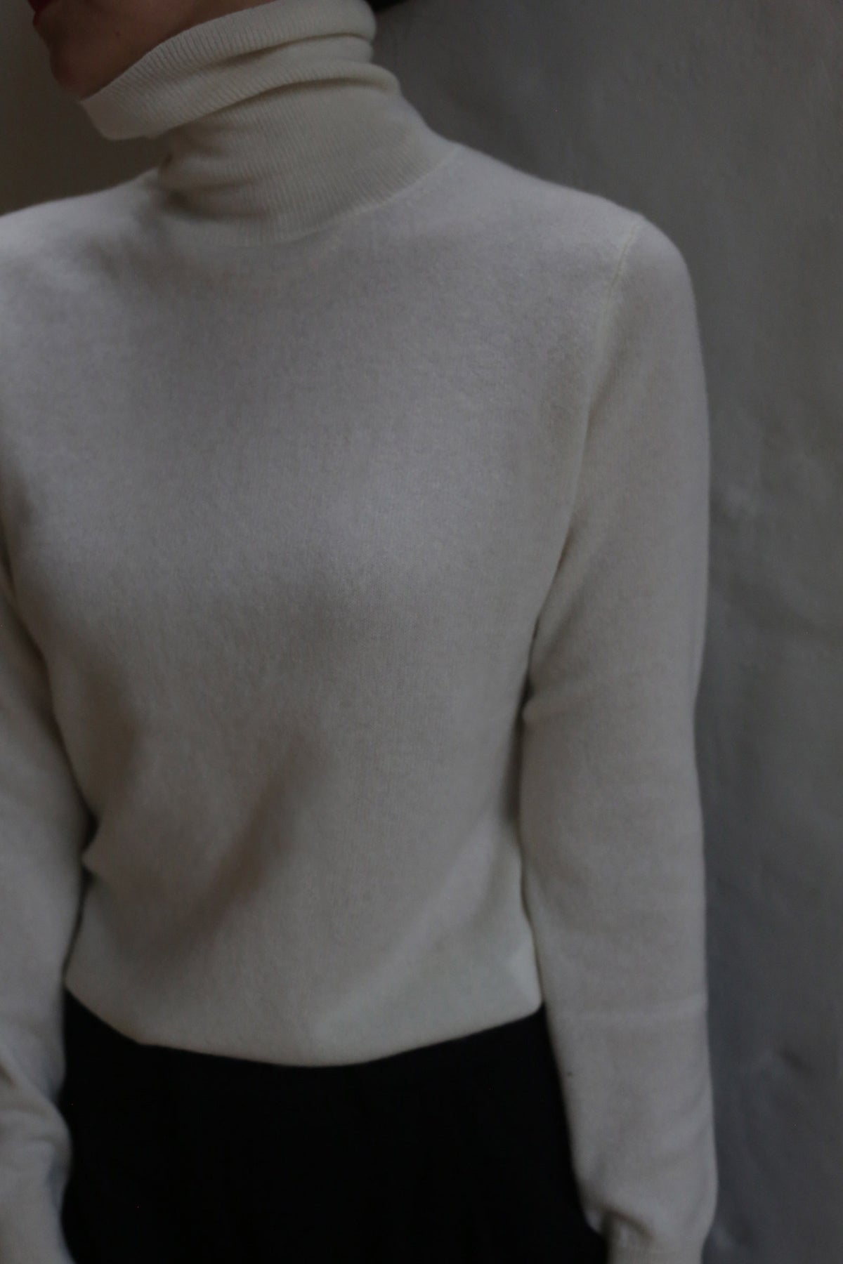 A person models the Ivory Cashmere Turtleneck against a neutral background, paired with dark pants. Their face is partially out of frame and mostly in shadow.