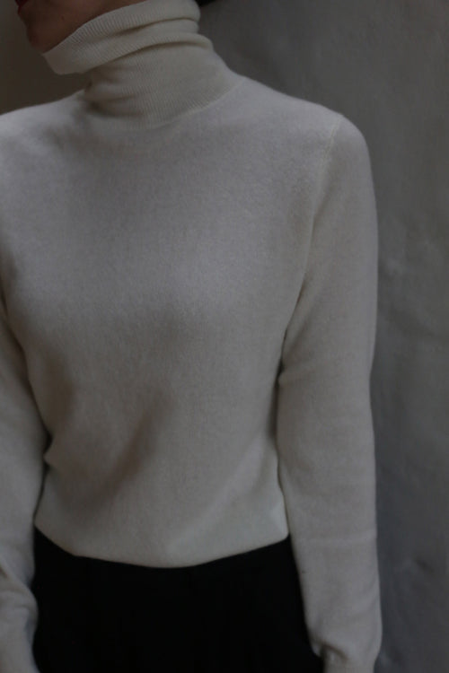 A person models the Ivory Cashmere Turtleneck against a neutral background, paired with dark pants. Their face is partially out of frame and mostly in shadow.