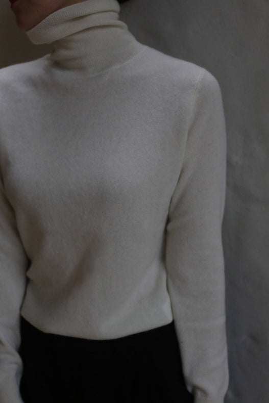 A person models the Ivory Cashmere Turtleneck against a neutral background, paired with dark pants. Their face is partially out of frame and mostly in shadow.