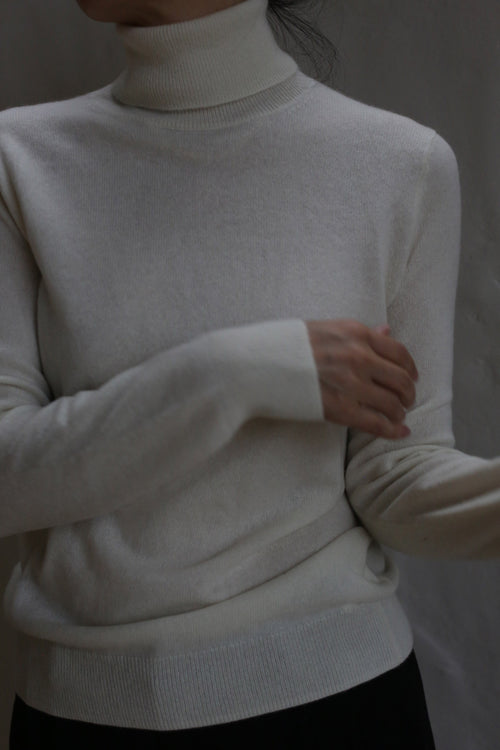 A person models the Cashmere Turtleneck in Ivory, standing against a neutral background with arms crossed at the waist and head partially out of frame.