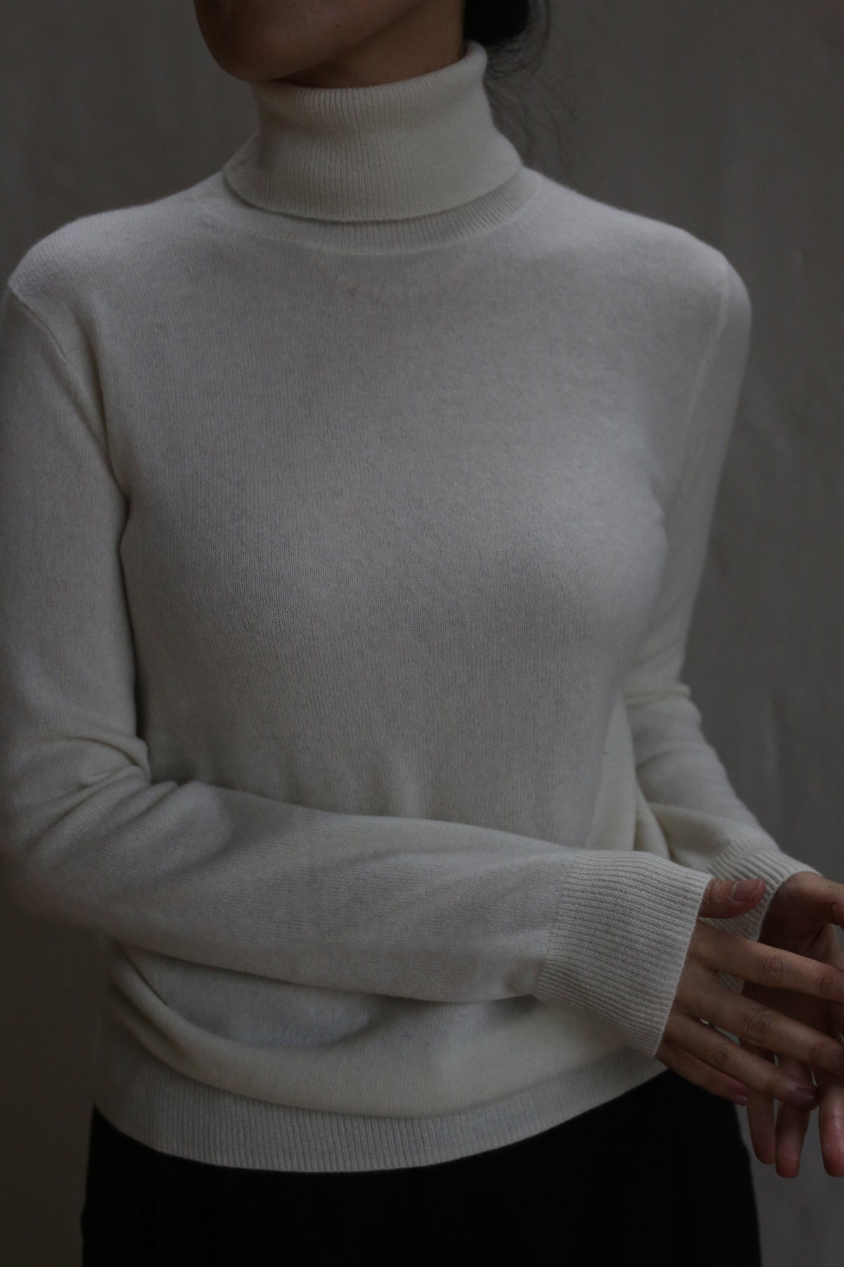 A person wearing the Cashmere Turtleneck in Ivory stands against a neutral background, their head partially out of frame and hands relaxed at the bottom right.
