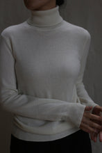 Load image into Gallery viewer, A person wearing the Cashmere Turtleneck in Ivory stands against a neutral background, their head partially out of frame and hands relaxed at the bottom right.