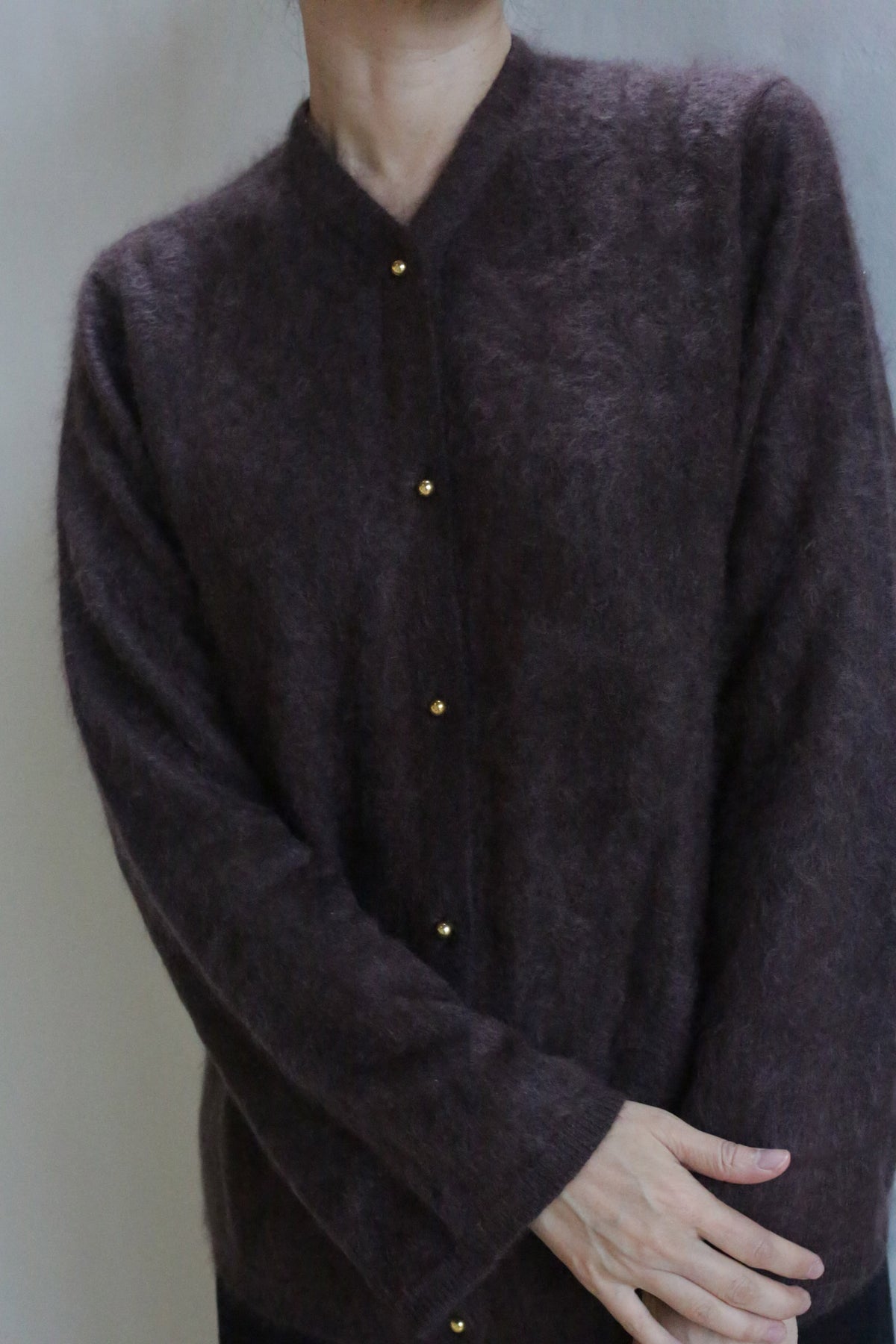 A person wears the Brushed Cashmere Cardigan in Mink, featuring brass buttons. They stand against a plain light background with arms crossed at the wrists and head slightly tilted down, partially out of frame.
