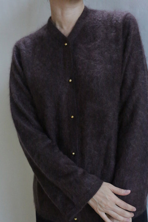 A person wears the Brushed Cashmere Cardigan in Mink, featuring brass buttons. They stand against a plain light background with arms crossed at the wrists and head slightly tilted down, partially out of frame.