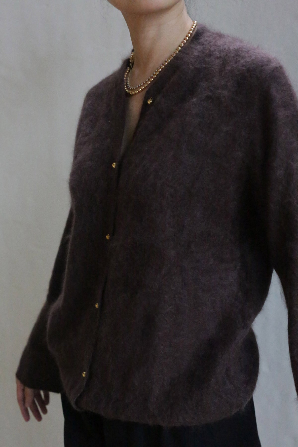 A person wears the Brushed Cashmere Cardigan | Mink—dark brown and fuzzy with brass buttons—and a gold beaded necklace, standing against a plain light background. Their head is partially out of frame.