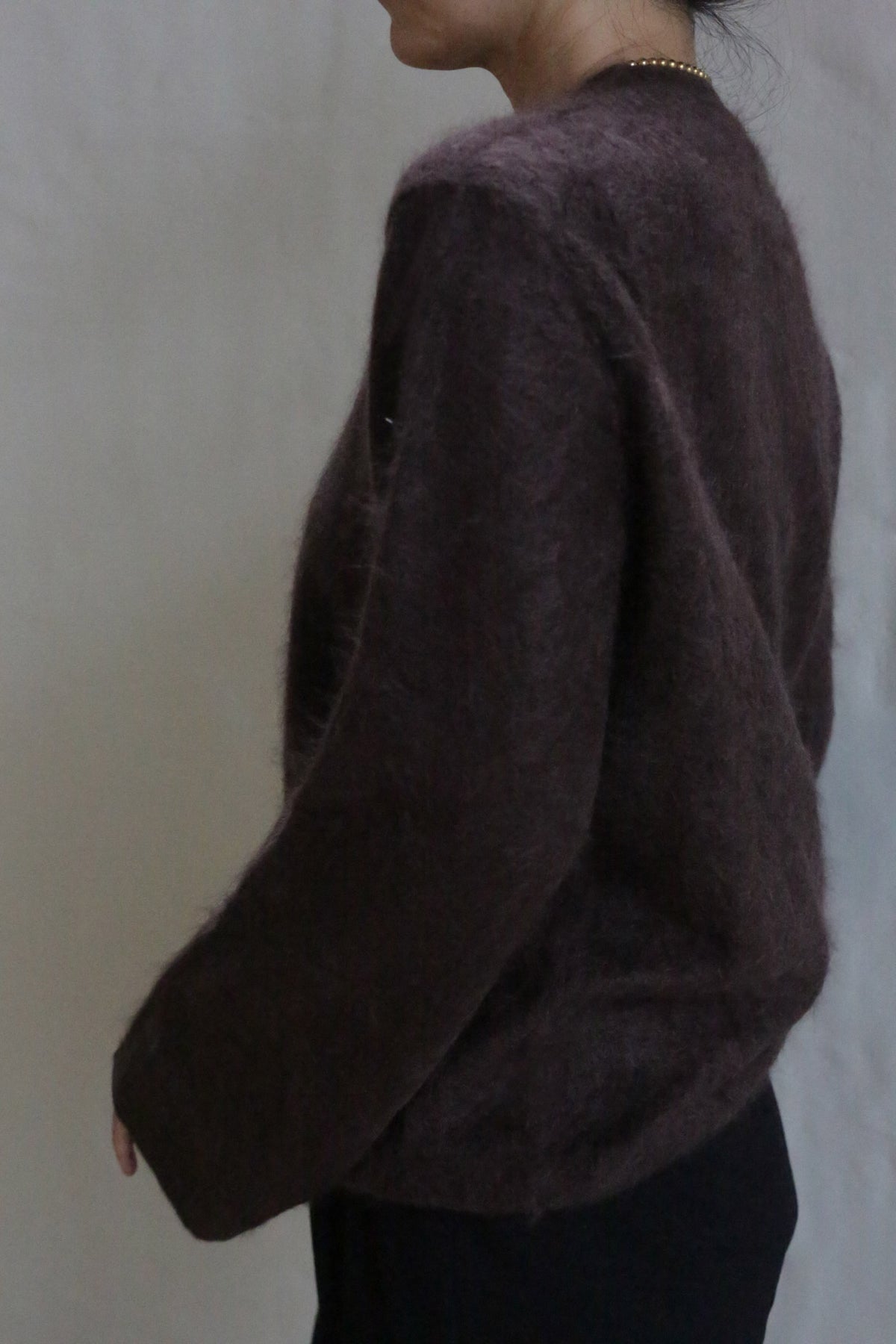 A person in the Brushed Cashmere Cardigan | Mink, which is loose and fuzzy in dark brown, stands sideways against a plain background, their head slightly turned away and a thin necklace visible.