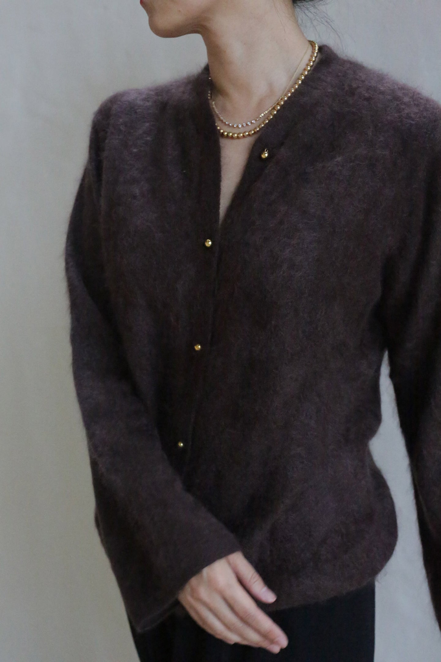 A person wearing the Brushed Cashmere Cardigan in Mink with brass buttons and a gold necklace stands against a plain background, their head turned to the side.