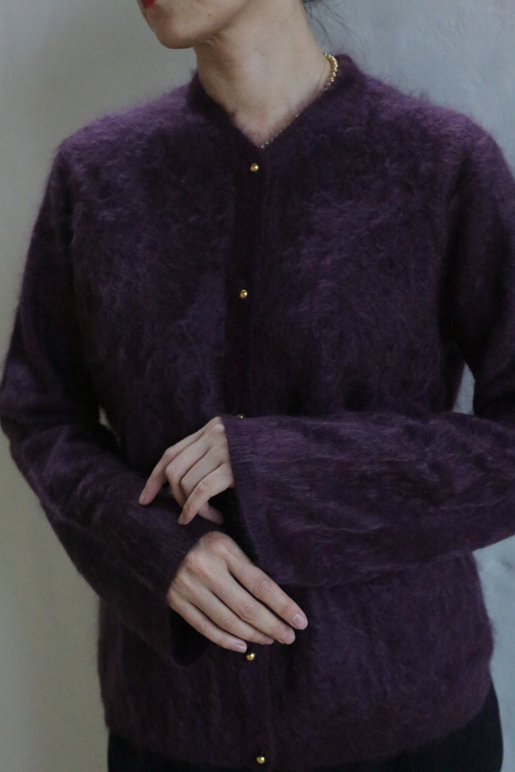 A person in a fuzzy Brushed Cashmere Cardigan | Aubergine with gold buttons stands against a neutral background, hands gently clasped. Their face is turned away, drawing focus to the cardigan’s luxurious, cozy texture.