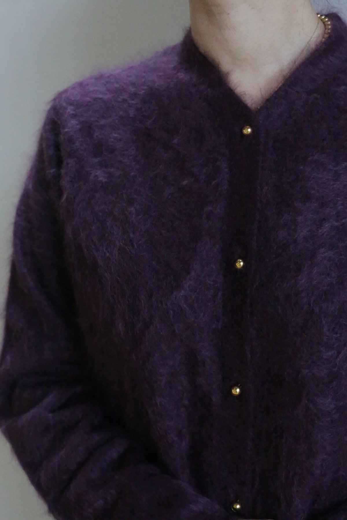 A person is shown from neck to waist wearing the Brushed Cashmere Cardigan in Aubergine, featuring gold buttons. The background is plain and the face is not visible, highlighting this luxury knitwear piece.