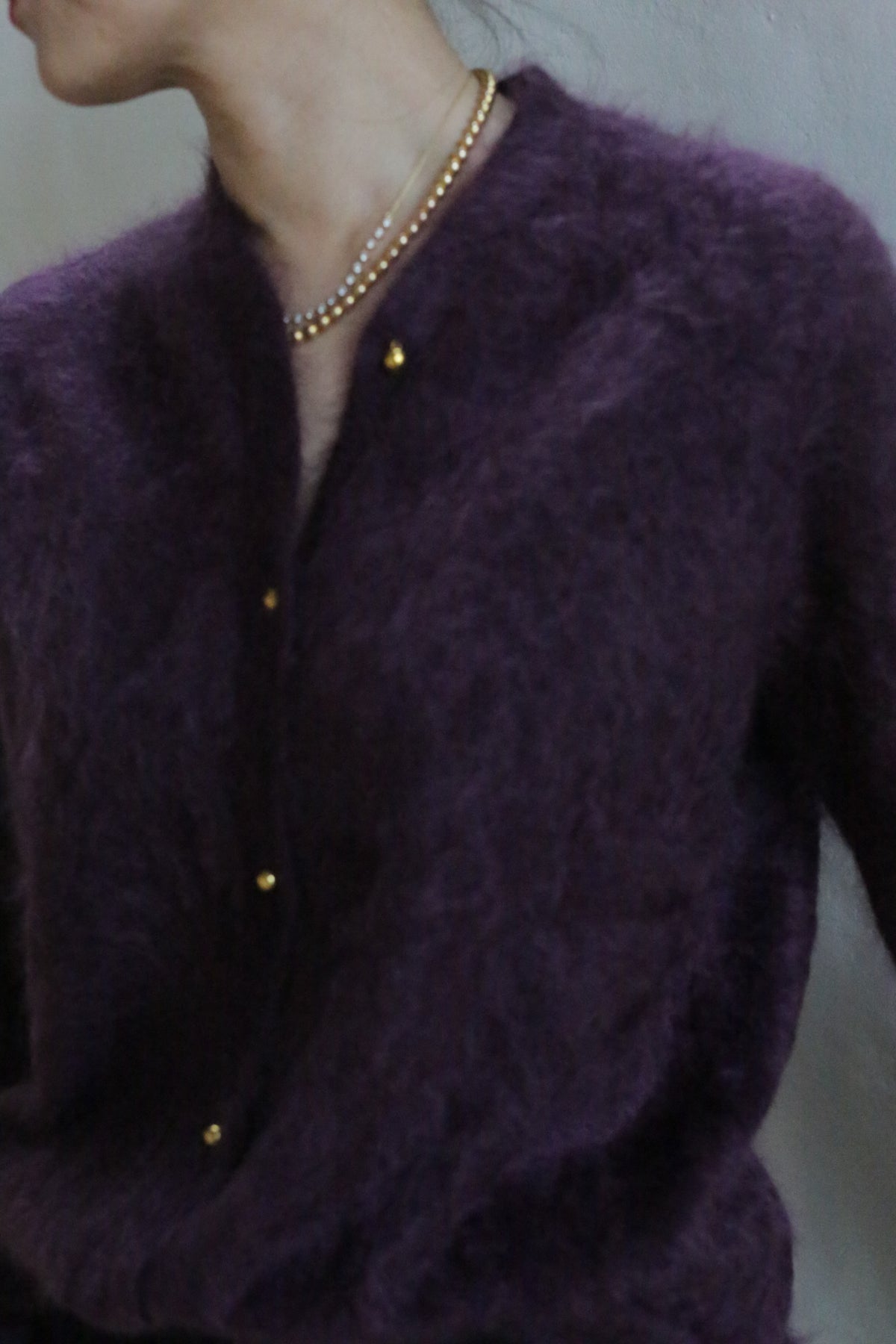 A person wearing the Brushed Cashmere Cardigan in Aubergine, featuring gold buttons and layered gold necklaces, shown from shoulders to waist, facing slightly away with their face partially out of view.