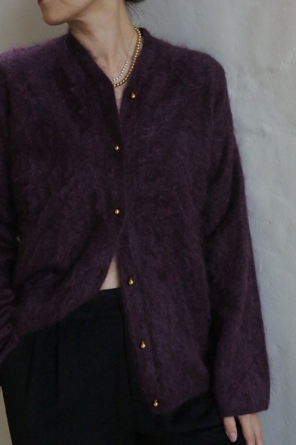 A person wearing the Brushed Cashmere Cardigan in Aubergine with gold buttons and black pants stands against a light wall, their head turned to the side, a pearl necklace visible—showcasing elegant luxury knitwear.