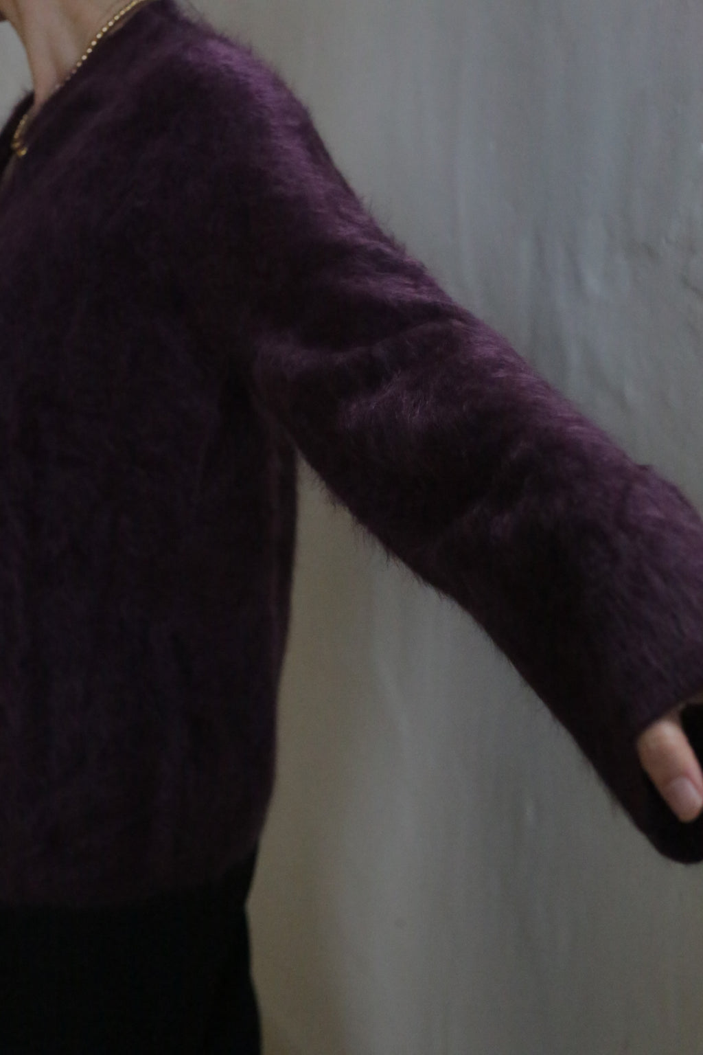 A person in the Brushed Cashmere Cardigan in Aubergine poses against a light-colored textured wall, showcasing only the upper body and outstretched arm.