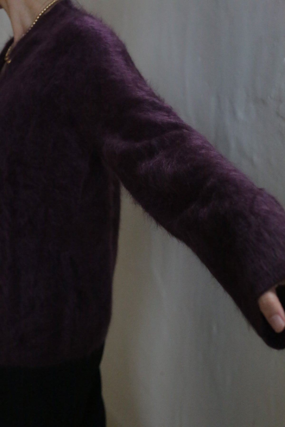 A person in the Brushed Cashmere Cardigan in Aubergine poses against a light-colored textured wall, showcasing only the upper body and outstretched arm.